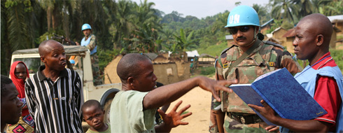 UN peacekeeper looks on as Community Liaison Assistant works in a field at Otobora, DRC.